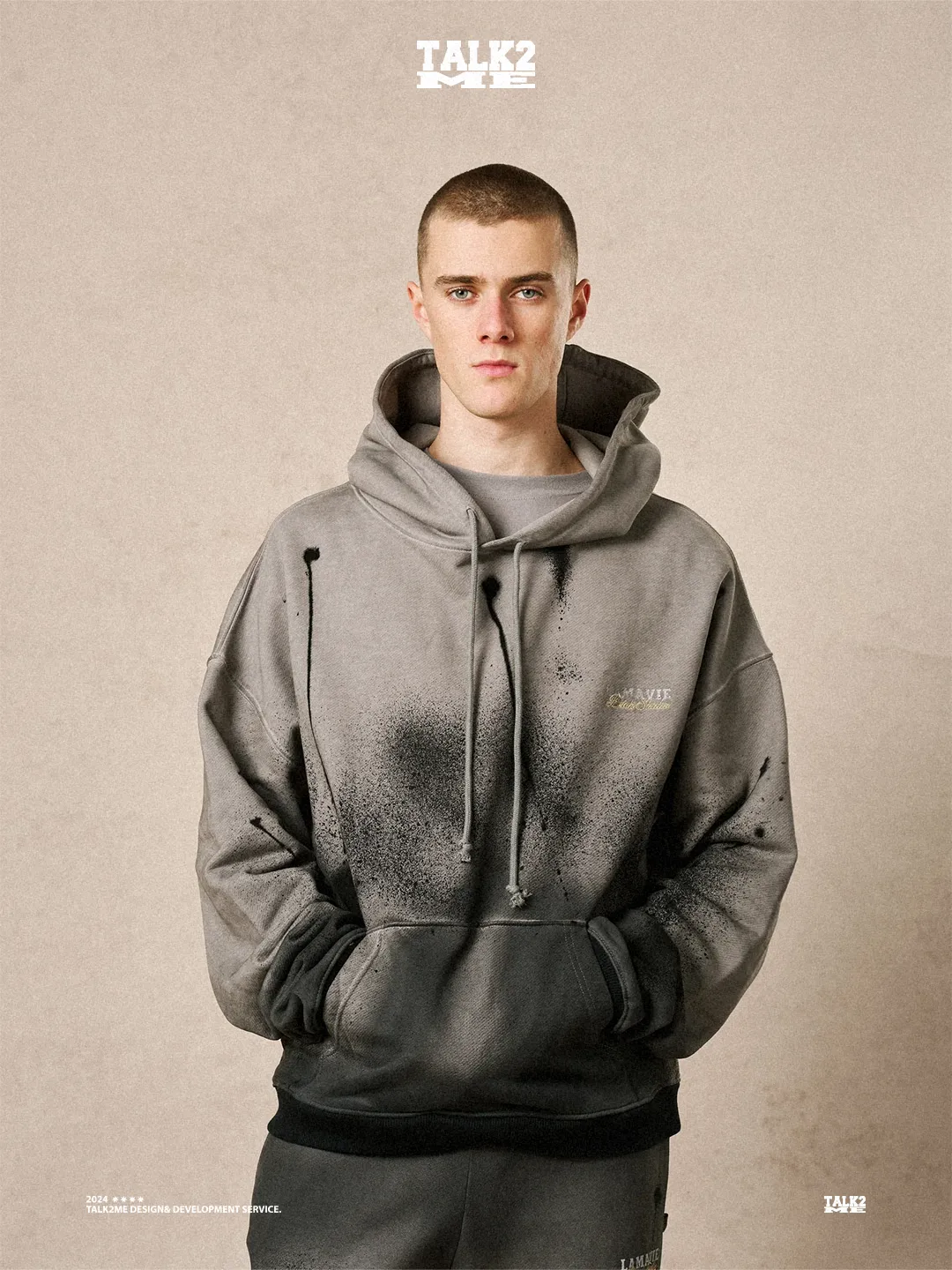 Content-Talks-Kampagne: Model im grauen La-Vie-Hoodie mit Sprenkel-Print, Porträt vor beiger Wand.