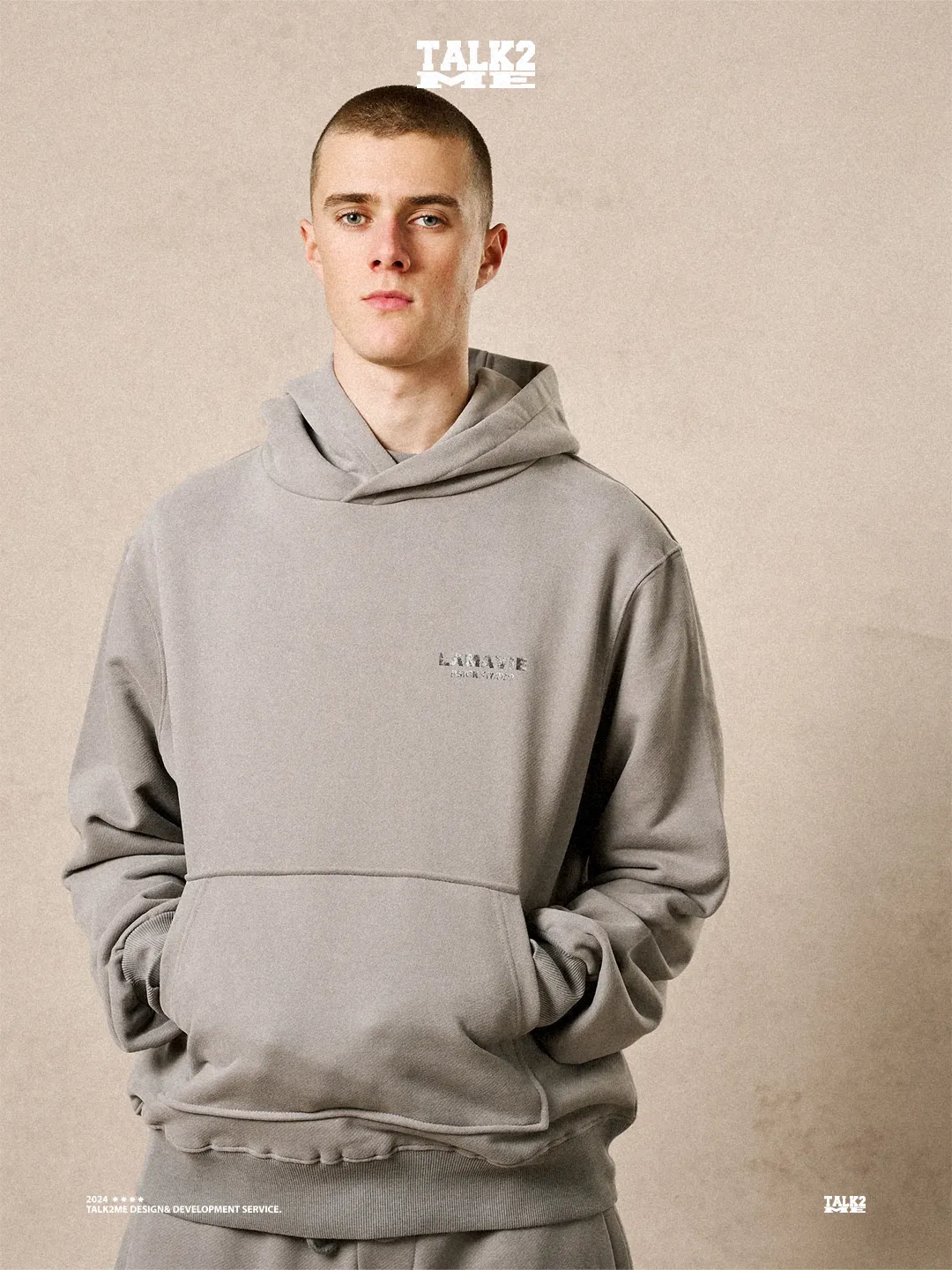 Content-Talks-Editorial: Model im grauen La-Vie-Hoodie, Porträt vor beiger Wand.